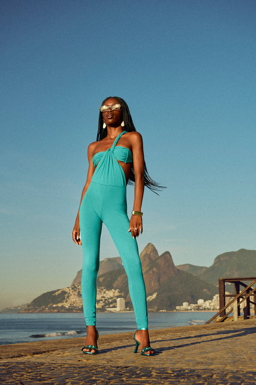 Model in a turquoise one-shoulder wrap jumpsuit with waist cut-outs, leaning against a palm tree on a tropical boardwalk.